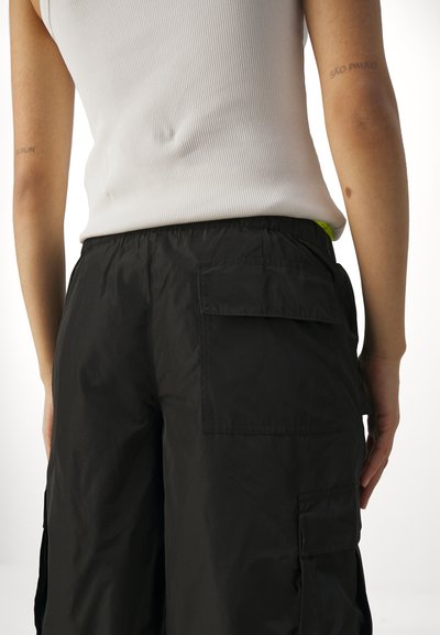 Zwarte cargo-broek met een achterzak, elastische tailleband en een gladde textuur. Gekleed met een aansluitend wit geribbeld tanktop.
