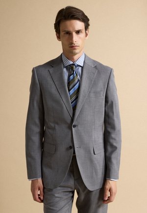 Man wearing a grey two-piece suit with a blue striped shirt and a striped green, blue, and black tie, standing against a beige background.