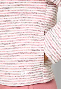 Gestreiftes Oberteil in Pink- und Weißtönen, mit gerippter Textur, seitlichen Schlitzen und einem Logoakzent am Saum. Lange Ärmel mit gestreiftem Design.