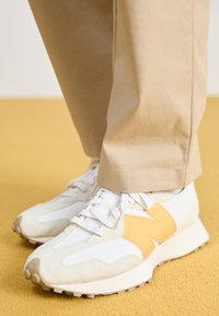 Weiße und beigetöne Sneaker mit einem gelben Akzent, strukturiertem Wildleder und einer Gummisohle mit markanten Profilen, kombiniert mit hellbeigen Hosen.