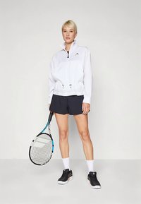 Vit vindjacka med dragsko i midjan, bärs över svart skjorta och shorts. Svarta tennisskor och en tennisracket i handen.