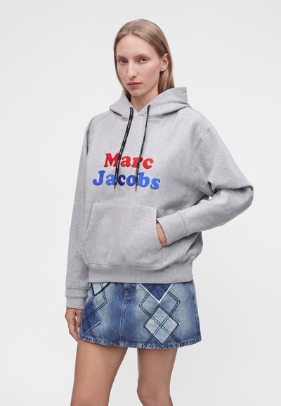Νεαρή γυναίκα φοράει γκρι φούτερ Marc Jacobs με κόκκινο και μπλε λογότυπο και μπλε τζιν φούστα με σχέδια διαμαντιού από patchwork.