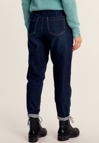 Jean en denim bleu foncé avec une coupe ajustée, présentant deux poches arrière et des ourlets retroussés. Porté avec des bottes à lacets noires et un pull en tricot.