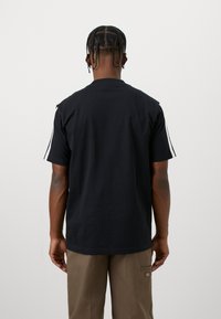 Άνδρας με πλεξούδες, φορώντας μαύρη μπλούζα με λευκές ρίγες στα μανίκια και καφέ παντελόνι, στραμμένος πλάτη σε απλό φόντο.