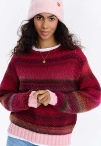 Sweter dziany w paski w odcieniach czerwieni i różu, z prążkowymi mankietami i dołem. Modelka ma na sobie dopasowaną różową czapkę beanie.