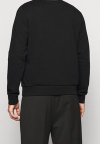 Sudadera negra con dobladillo y puños acanalados, que presenta un pequeño logo dorado en el cuello y una tela suave y texturizada.