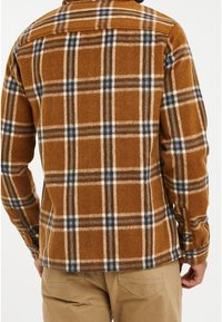 Mand iført en brune, blå og hvide ternede flannel skjorte med beige bukser, vist fra ryggen mod en lys hvid baggrund.
