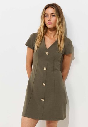 Frau mit langen blonden Haaren, die ein kurzärmeliges olivgrünes Kleid mit vier großen goldenen Knöpfen vorne trägt.