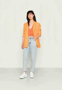 Blazer laranja sobre uma blusa laranja, combinado com jeans de lavagem clara e ténis brancos. Tecidos suaves, corte descontraído e design em camadas.