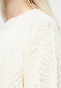 Κοντινή λήψη ενός ατόμου που φοράει λευκό πουλόβερ με πλεκτό σχέδιο, εμφανίζοντας λεπτομερή υφή στους ώμο και το πάνω μέρος του χεριού.
