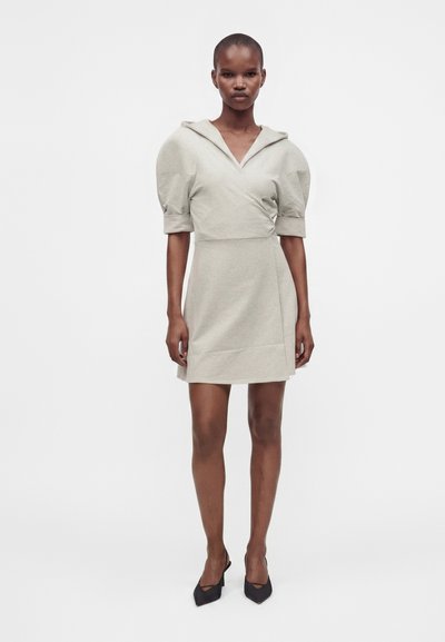 Femme portant une robe beige clair avec des manches courtes bouffantes, un profond décolleté en V et des talons noirs pointus, debout devant un fond uni.