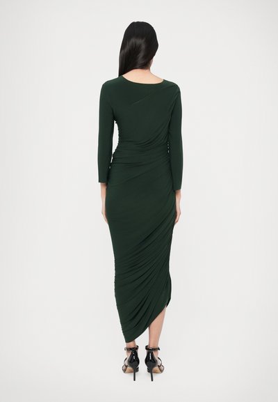 Ciemnozielona sukienka z długim rękawem z marszczonymi draperiami z tyłu, tworząca smukłą sylwetkę. Modelka ma na sobie czarne sandały na wysokim obcasie.