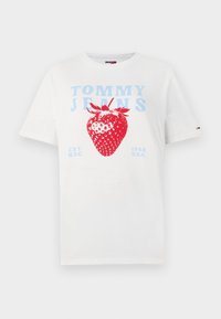 T-shirt en coton blanc avec un grand motif de fraise rouge et le texte bleu "TOMMY JEANS". Design simple, manches courtes, col rond.