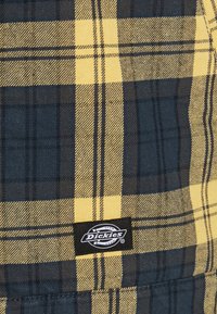 Černá, žlutá a tmavě modrá károvaná tkanina s texturou připomínající tkanou bavlnu. Na spodním pravém rohu se nachází nášivka s logem Dickies.