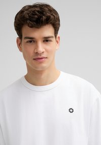 Witte katoenen t-shirt met een ronde hals en korte mouwen, voorzien van een klein zwart logo op de linker borst. Gladde textuur, casual pasvorm.
