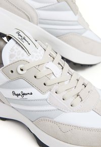 Balti ir beige sportbačiai, pagaminti iš lygaus ir tekstūruoto medžiagų derinio, su logotipu ir akcento juostomis, apvalia nosimi ir raištelių uždarymu.