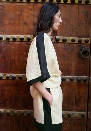 Kobieta w beżowym stroju z szerokimi czarnymi paskami na rękawach i szortach stoi bokiem przy drewnianych drzwiach z metalowymi ćwiekami i okrągłą klamką.