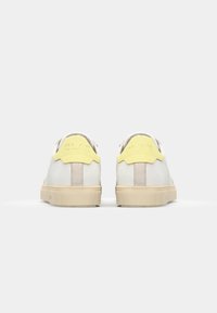 White suede sneakers with yellow and beige accents, featuring a textured, cream rubber sole, and a CLAE logo on the back.