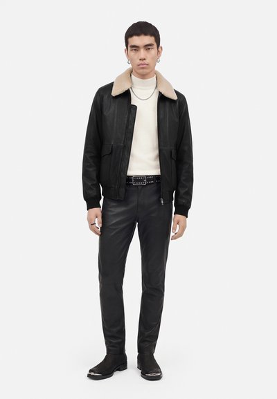 Jeune homme portant une veste en cuir noire avec col en peau lainée beige, un col roulé blanc, un pantalon en cuir noir, une ceinture cloutée et des bottes noires.
