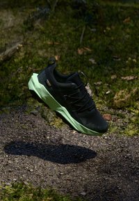 Fekete és zöld Gore-Tex terepfutó cipő egy kavicsos ösvény felett lebeg, a háttérben fű és moha látható.