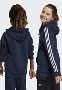 Sudadera con capucha de color azul marino, con tres franjas blancas a lo largo de las mangas, de tejido suave, de mangas largas y corte relajado, llevada por dos niños.