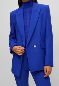 Niebieska, dopasowana marynarka z szpiczastymi klapami, pojedynczym srebrnym guzikiem i gładką fakturą. Noszona na dopasowanym, prążkowanym topie z wysokim kołnierzem.