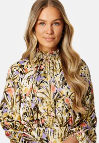 Blouse florale en tissu léger, avec un col froncé haut et des manches longues. Le motif comprend des fleurs vertes, violettes et roses sur une base noire.