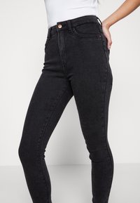 Vaqueros skinny negros de denim, con una cintura alta, ajuste ceñido y detalle de botón de cobre en la cinturilla. Textura suave con mínimas desgastes.