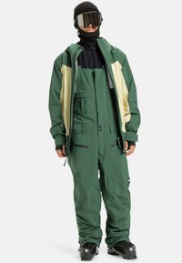 Persoon in een groen en crèmekleurig sneeuwjack en groene bibbroek, met een zwarte beanie, skibril, zwarte handschoenen en skischoenen op een witte achtergrond.