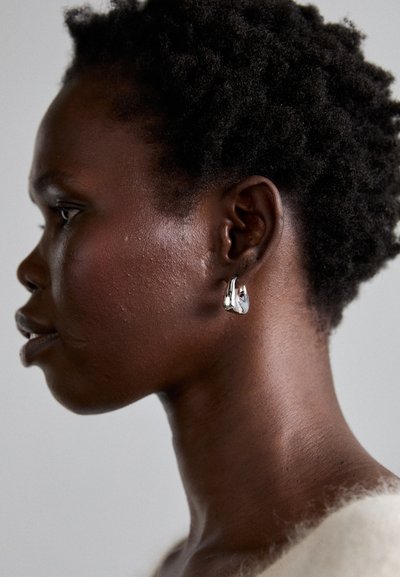 Silver hoop earring on textured skin, showcasing a sleek, modern design with curves and a polished finish, set against a light background.