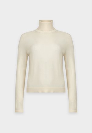 Femme portant un pull à col roulé crème, un pantalon marron à jambes larges, des chaussures beige pointues et des boucles d'oreilles créoles dorées, debout devant un fond uni.