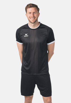 Uomo in piedi sorridente che indossa una maglietta sportiva nera con rifiniture bianche e pantaloncini neri abbinati su uno sfondo semplice.