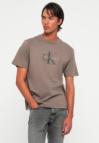 Man with medium skin tone wearing taupe Calvin Klein t-shirt and gray jeans, standing with one hand in pocket, looking right.