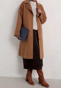 Brown wool coat with large lapels, worn over a light gray knitted sweater. Dark brown corduroy skirt and brown heeled boots. Blue clutch bag.