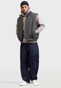 Melnā un pelēkā rakstā izšūta polsterēta veste uz gaiši pelēka kapuču džempera, kombinēta ar tumši zilām platām džinsa biksēm un baltām sporta kurpēm.