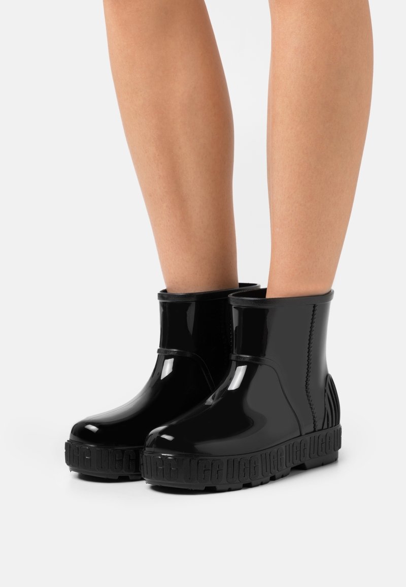 Bottes noires brillantes pour la cheville avec semelles épaisses embossées portées sur des jambes nues contre un fond clair uni.
