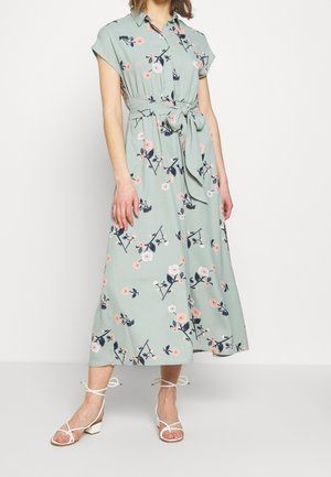 Femme portant une robe midi verte à fleurs avec manches courtes, col chemise, ceinture à la taille et sandales blanches à brides et talons bas.