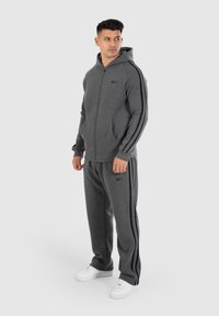Haut à capuche zippé gris avec des rayures noires sur les côtés, assorti à un pantalon de survêtement. Des baskets blanches complètent la tenue, mettant en avant un design décontracté.