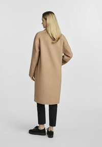 Długi, beżowy płaszcz z wełny z prostym krojem, opuszczonymi ramionami i kieszeniami naszytymi. Noszony na czarnych spodniach i ciemnych butach.