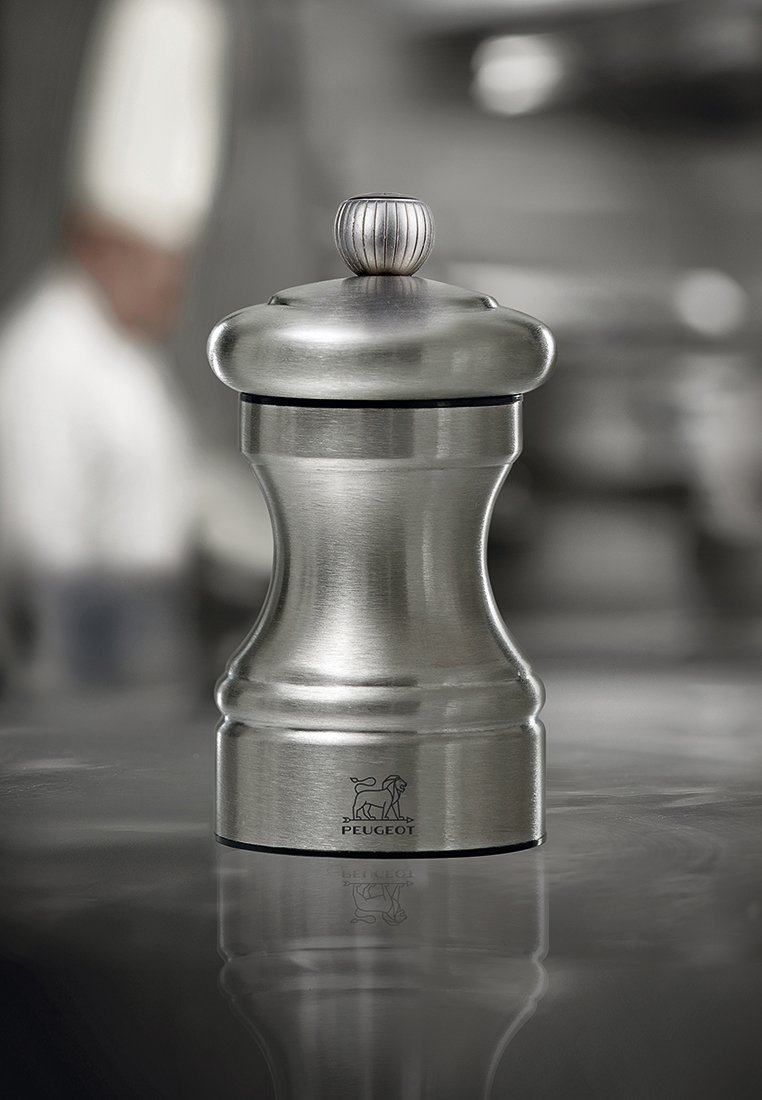 Edelstahl-Pfeffermühle von Peugeot, zentriert auf einer reflektierenden Oberfläche, mit unscharfer Kochfigur in weißer Uniform im Hintergrund.