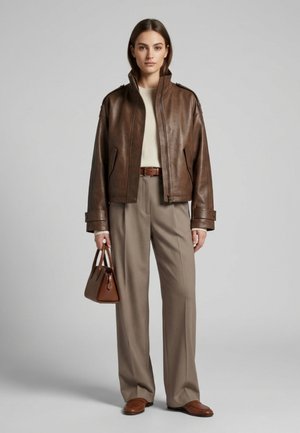 Frau trägt eine braune Lederjacke, einen beigen Rollkragenpullover, beige Hosen mit weitem Bein, einen braunen Gürtel, braune Loafers und hält eine braune Handtasche.