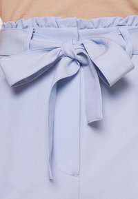 Ljusblå högmidjade byxor med ett tygbälte knutet i en rosett, bärs tillsammans med en beige ribbad topp som är instoppad.