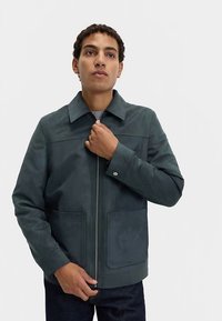 Dunkelgrüne Jacke mit Reißverschluss, breitem Kragen, zwei Fronttaschen und glatter Textur. Verfügt über minimale Nähte und Knopfakzente.