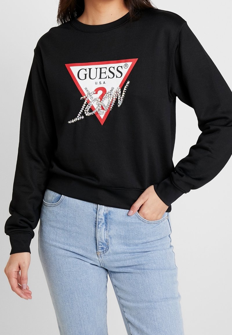 Felpa nera con un logo triangolare rosso recante la scritta "GUESS U.S.A." e dettagli simili a diamanti sul davanti, abbinata a jeans blu chiaro.