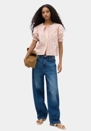 Kvinde iført en pink kortærmet bluse, blå jeans med vide ben, hvide sandaler, der holder en foldet brun jakke mod en ensfarvet baggrund.