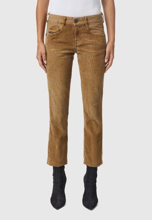 Jasnobrązowe spodnie z sztruksu o prostych nogawkach, noszone z czarnymi botkami na obcasie z ostrymi noskami, na osobie z widocznymi tatuażami na ramionach i białym topem.