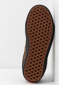 Semelle en caoutchouc d'une chaussure noire avec une bande de roulement marron présentant des motifs hexagonaux et diamant, positionnée verticalement sur un fond blanc.