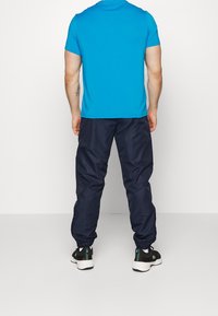 Man som bär en ljusblå kortärmad skjorta, mörknavy joggerbyxor, vita strumpor och svarta sneakers, stående vänd bort.