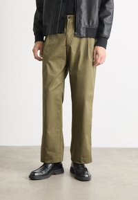 Pantalons larges vert olive en tissu lisse, associés à des chaussures noires. La tenue présente un design simple et épuré, sans motifs visibles.