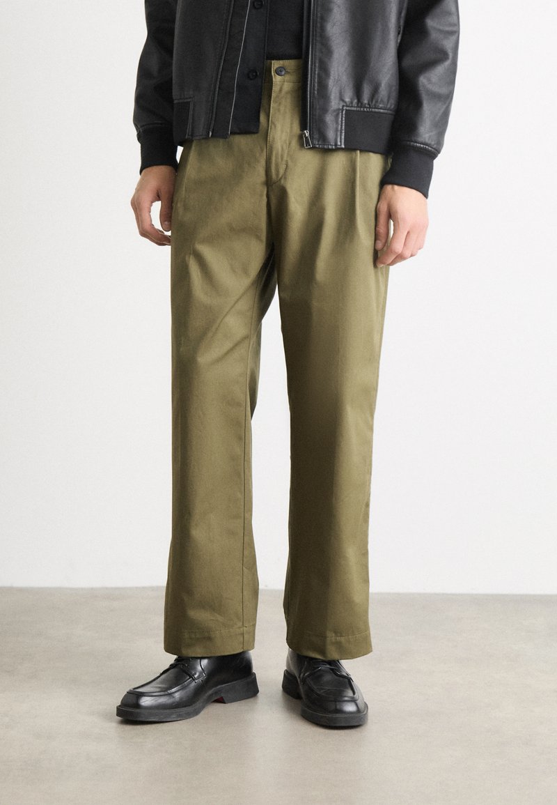 Pantalons larges vert olive en tissu lisse, associés à des chaussures noires. La tenue présente un design simple et épuré, sans motifs visibles.